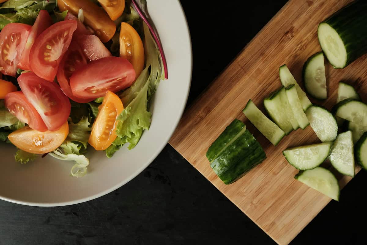 A bowl of lettuce with tomatoes on top and a sliced cucumber on a cutting board